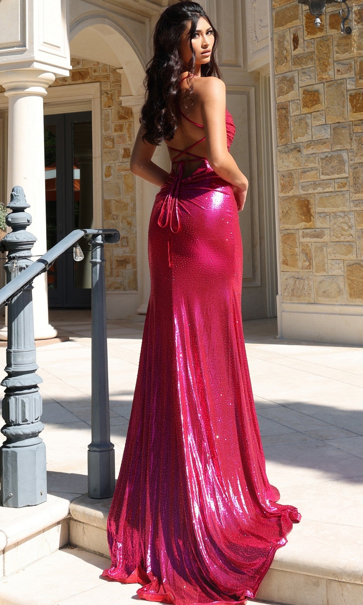 Wearing the Jovani 47752 long prom dress in shiny bright pink, a woman stands on outdoor steps. The fitted, floor-length gown features crisscross back straps for an elegant touch as she looks over her shoulder.